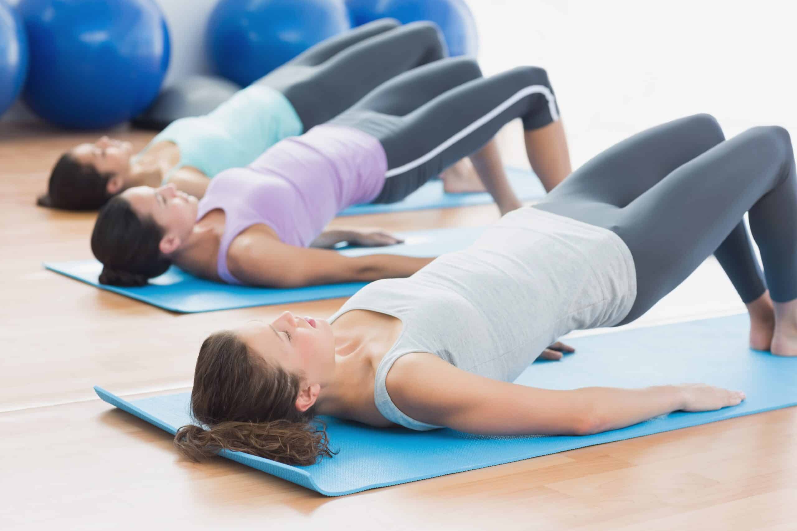 Trois femmes exécutent l'exercice du pont sur des tapis bleus lors d'un cours de pilates ou de fitness en salle. Elles renforcent le dos.